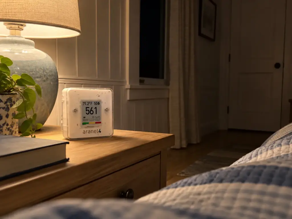 Compact CO2 monitor on a coastal cottage bedroom nightstand beside a lamp, the room dark around it
