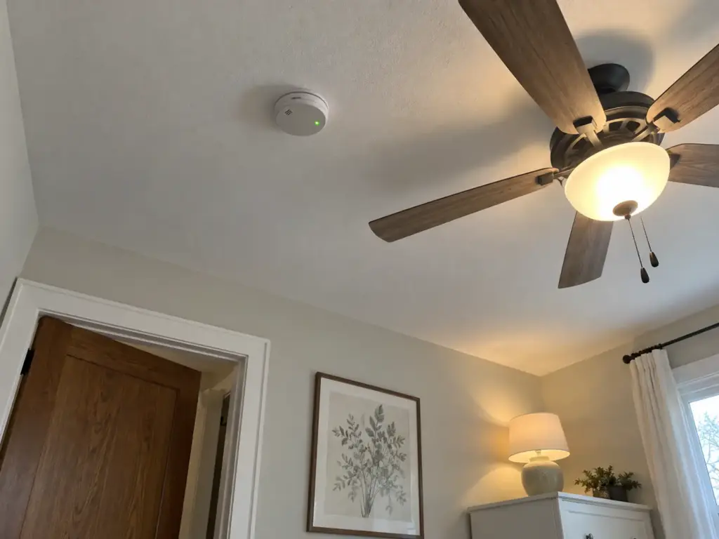 Smoke and carbon monoxide alarm on a bedroom ceiling beside a ceiling fan with its light on